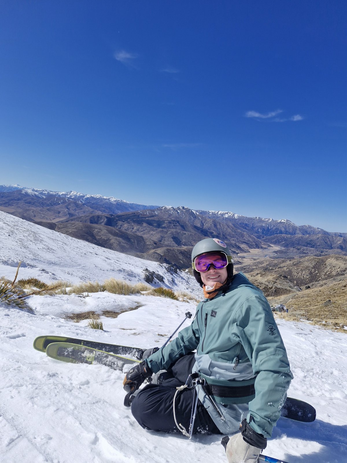 Noah sitting on a snowy slope in ski gear, mountains behind