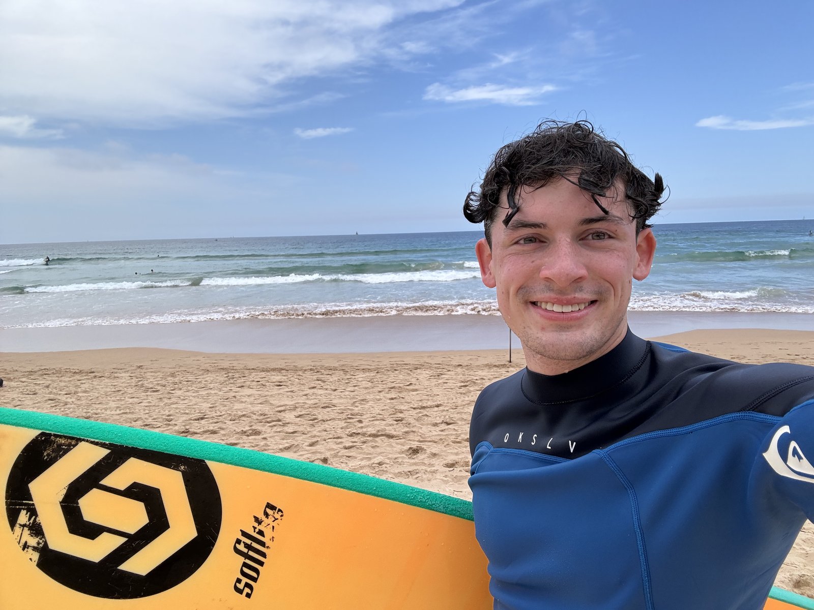 Noah at the beach in a wetsuit with his surfboard