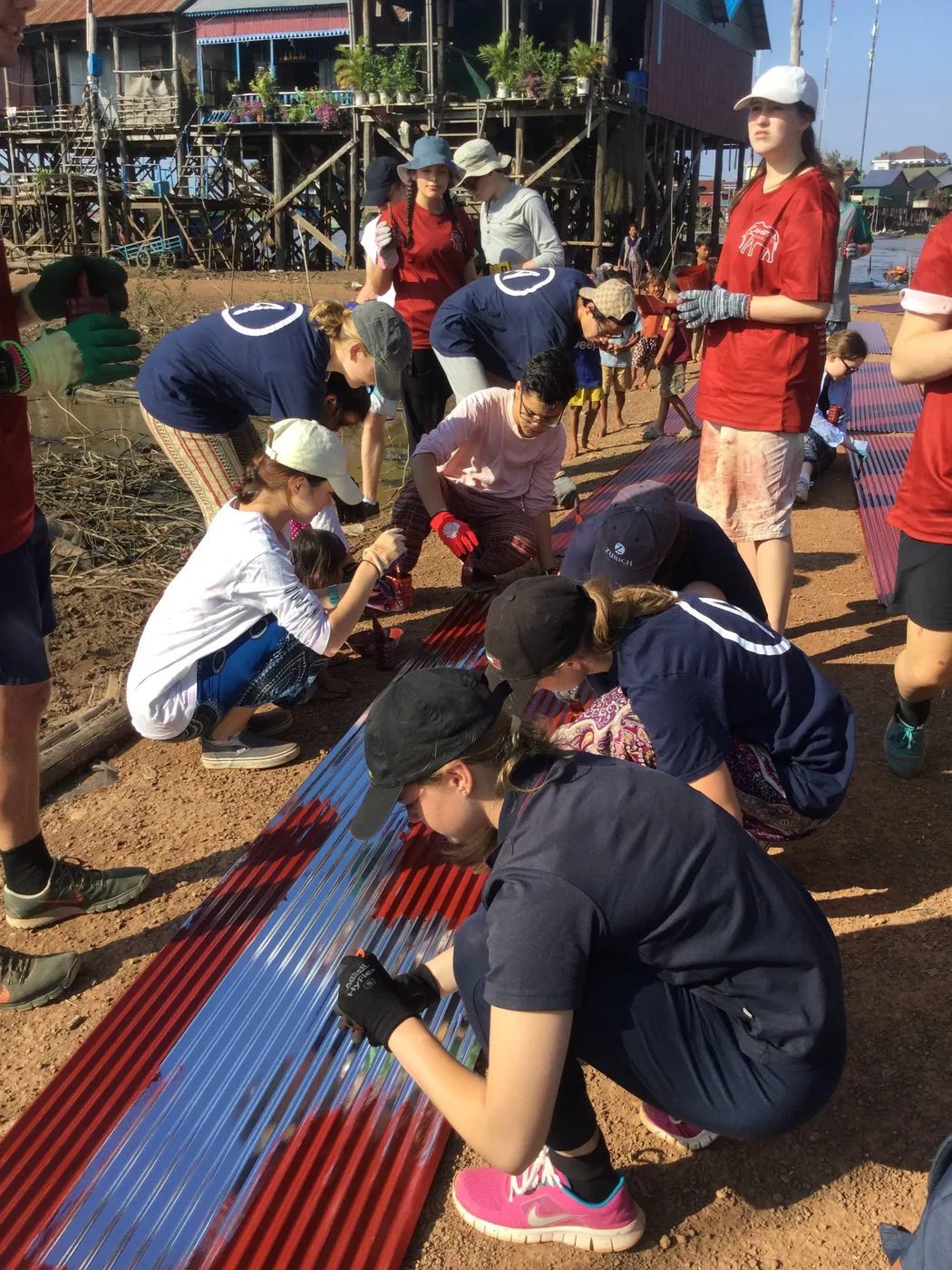 Volunteers kneeling on red earth painting corrugated roofing sheets beside a Cambodian stilt village